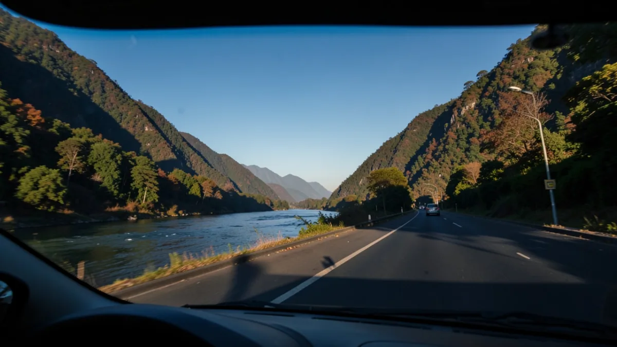 A view from a car window showing the Teesta River flowing alongside a mountain road, with green hills under a clear blue sky.